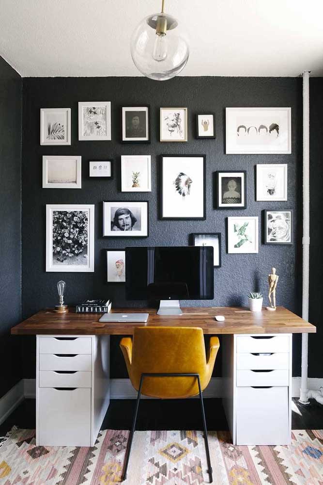 Home office decorado com quadros e fotos sobre a parede escura; repare que a mesa foi feita com uma tábua de madeira apoiada sobre dois gaveteiros