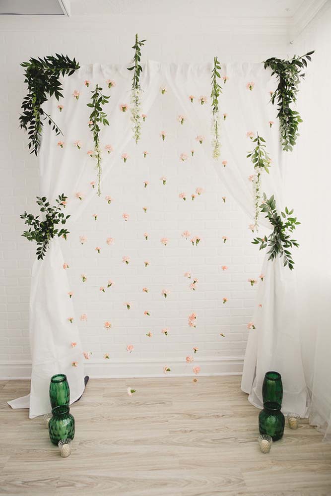Espaço para fotos super romântico com arco decorado com cortina de TNT branco, aplicação de folhas e uma cascata de flores.