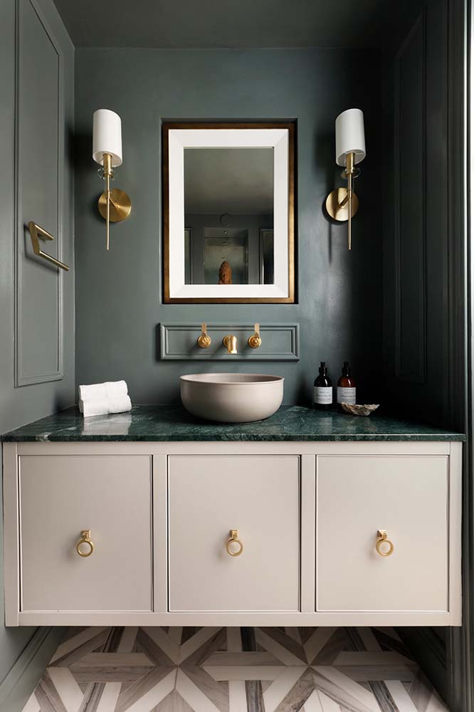 A pintura verde musgo intenso nas paredes cria uma unidade com a bancada de granito e contrasta com os armários beges e os metais dourados desse lavatório elegante.