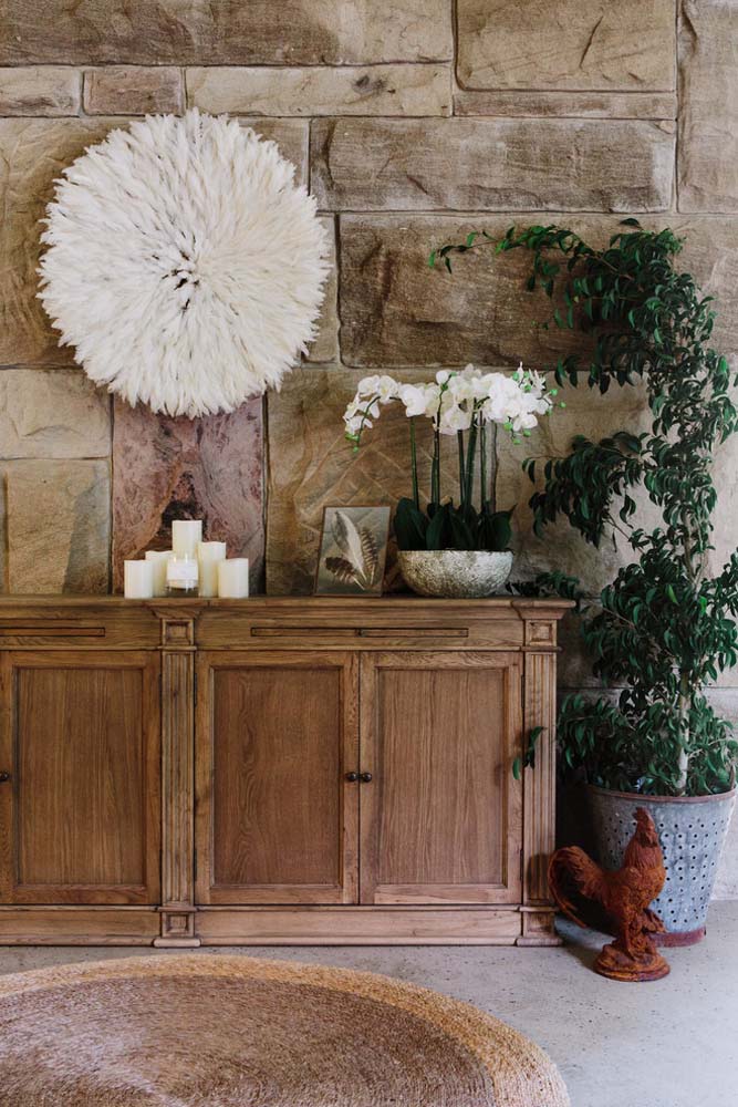 Rústico, elegante e todo inspirado na natureza: um espaço com revestimento de pedra na parede, cômoda de madeira e vasos de planta. 