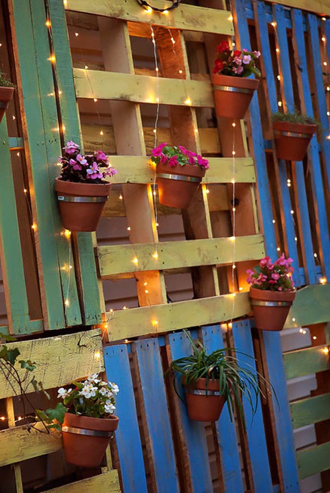 Um visual criativo e aconchegante para a área externa, com floreira de pallets dispostos na parede intercalando o direcionamento e as cores, com vasinhos de cerâmica e cordão de luzes. 