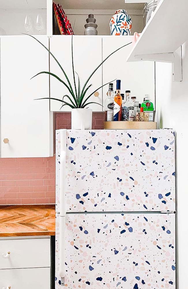 Com toques de cor e aparência vintage, a geladeira envelopada com estampa de granilite ainda abriga uma bandeja-bar e um vaso de planta acima dela. 