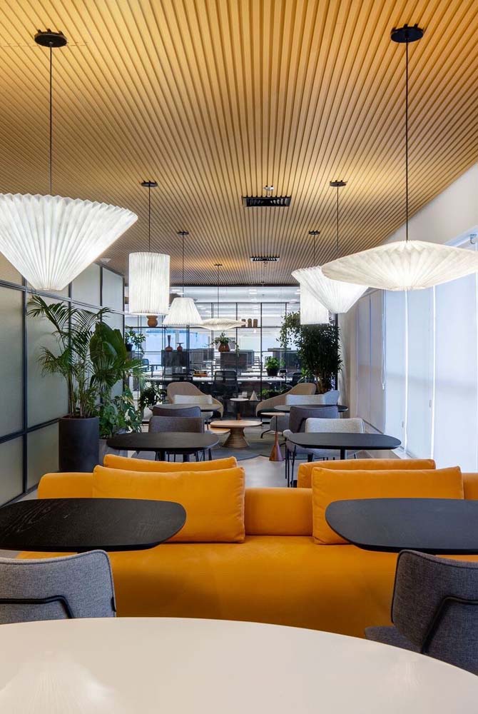 O sofá ilha mostarda substitui as cadeiras de um lado das mesas nesta área para refeições e descanso no escritório. 
