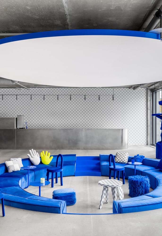 Moderno e cheio de personalidade, esse espaço coletivo no escritório é composto por dois sofás semi circulares azuis em um desnível no piso. 